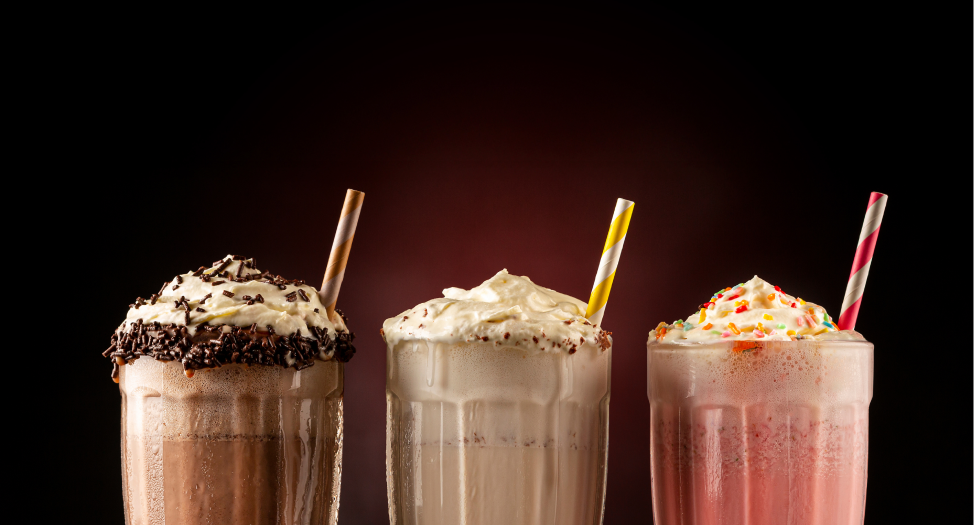 Three milkshakes with whipped cream and straws, featuring chocolate, vanilla, and strawberry flavors.
