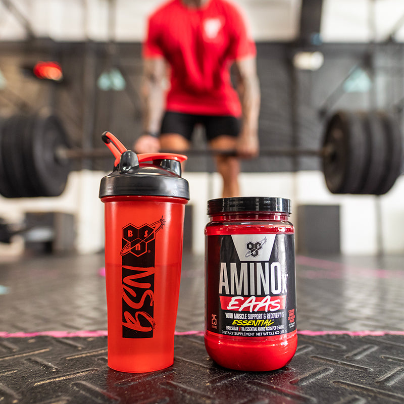 BSN shaker bottle and Amino X EAAs supplement container on gym floor with weightlifter in background.