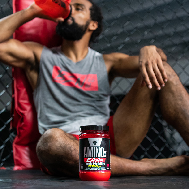 BSN Amino X EAAs supplement container with a person drinking from a shaker bottle in a gym setting.